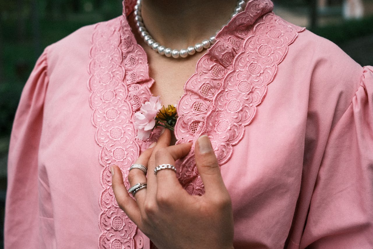 A woman's hand holds a flower against a pink lace blouse, featuring elegant pink attire and pearl necklace.